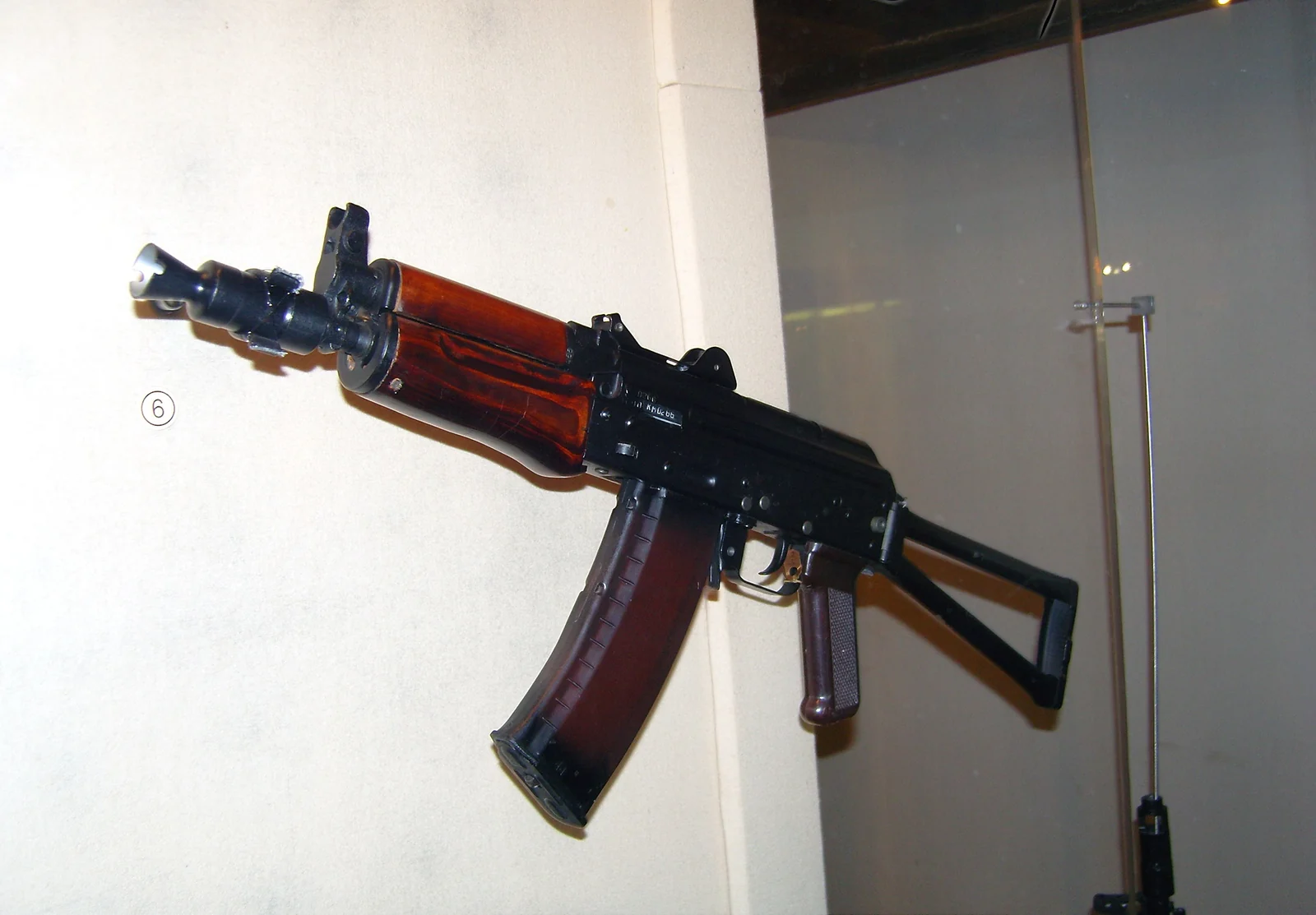 A short black carbine with a wooden pistol grip, a curved orange-tinted magazine and a folding metal stock, photographed at an angle against a plain wall.