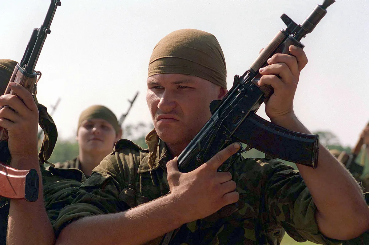 A soldier in a bandana holds a short carbine upright while standing in a line with other soldiers.