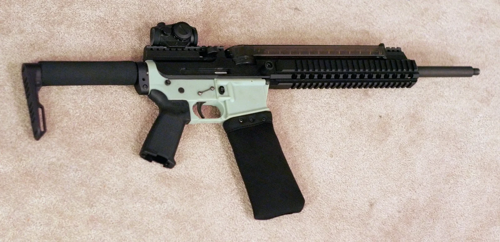 An AR-15-pattern carbine with a horizontal magazine sheathed in a black cloth cover running above the handguard, a red-dot sight forward of a light green lower receiver, and a fixed carbine-length stock.