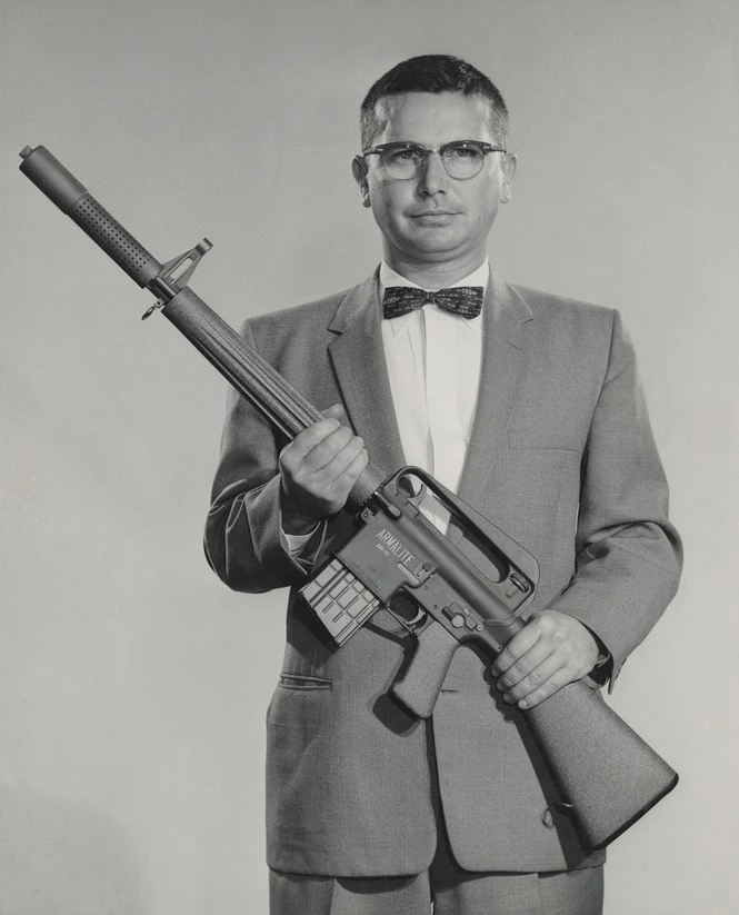 A man in a suit and bow tie stands holding an early rifle prototype with a fixed bayonet, its stock and furniture visibly different in shape from later production AR-10s.