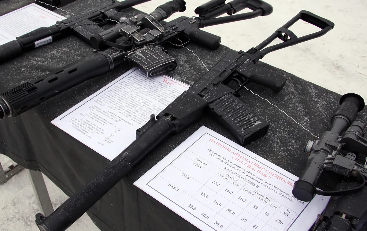 Three suppressed Russian weapons laid on a snow-dusted black display table with Russian-language specification cards between them. The nearest carries a large telescopic sight; the middle one has a folding wire stock and a ribbed magazine; a printed table lists SP-5, SP-6 and PAB-9 cartridge data.