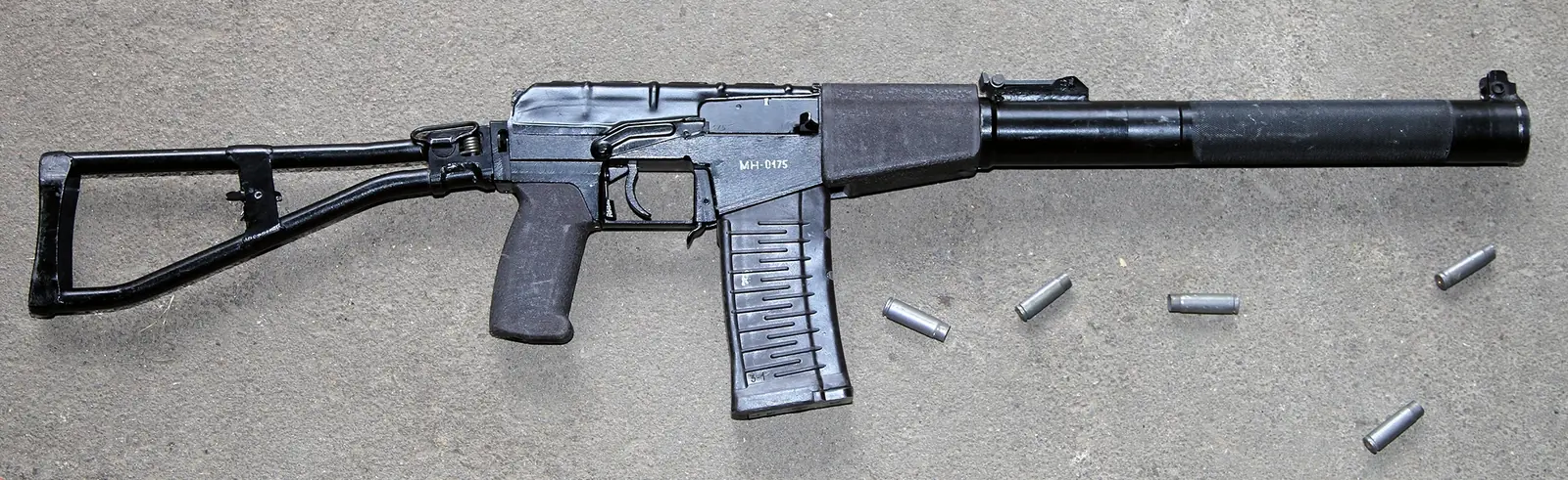 A black Kalashnikov-pattern rifle lying on concrete, seen from the right with a tubular skeleton stock extended to the rear. Almost the whole front half of the weapon is a fat cylindrical casing running from the receiver to the muzzle, wider than the receiver itself, with a front sight on top of it. A short curved magazine with horizontal ribs sits below. Several fired cases lie beside it.