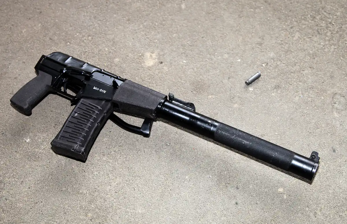 The same rifle on concrete with its wire stock folded flat along the left side of the receiver, so the weapon ends immediately behind the pistol grip. The suppressor tube still makes up more than half its total length.