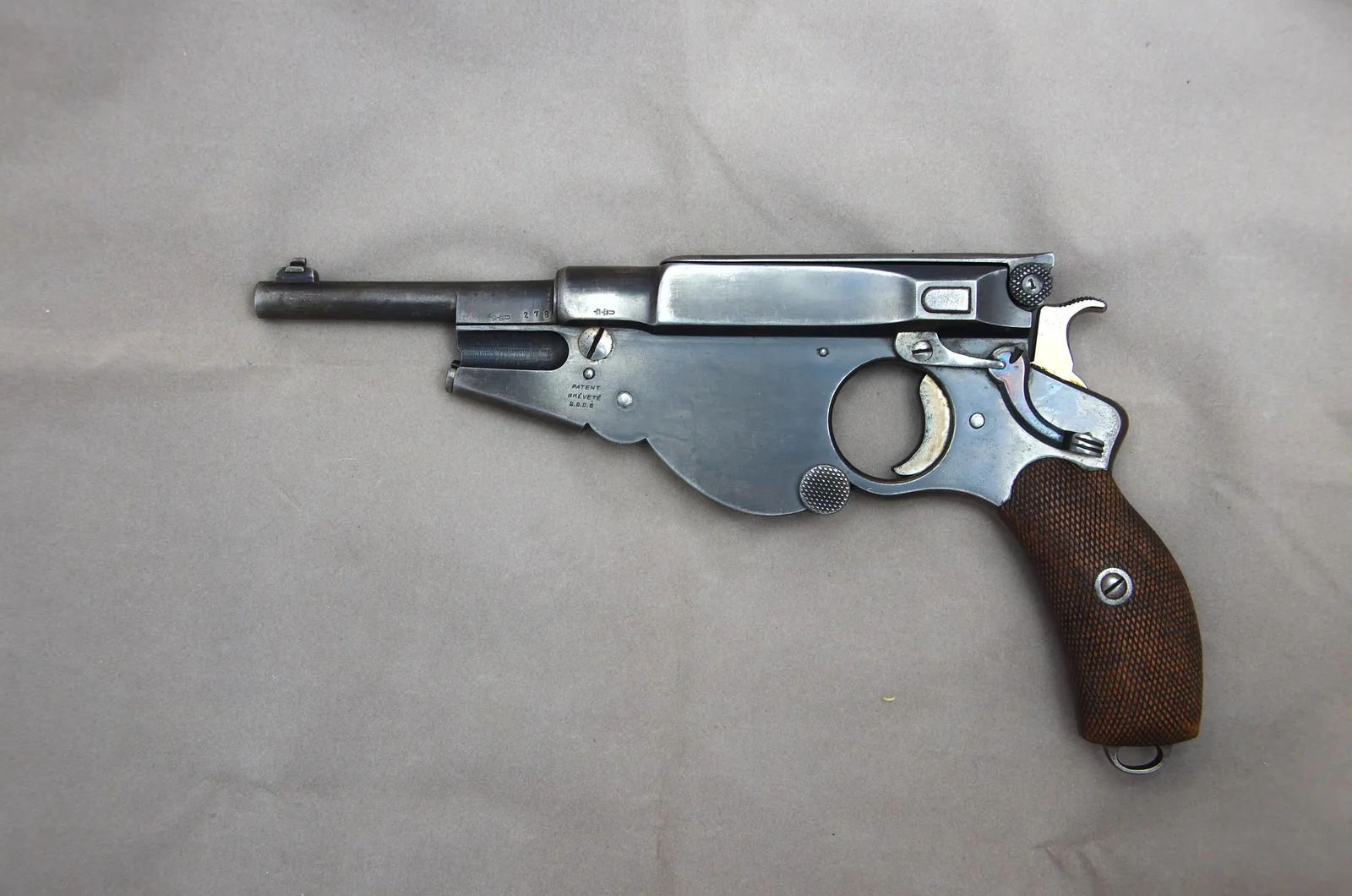 A small early automatic pistol with a wooden grip, a short round barrel and an exposed hammer, photographed from the left.