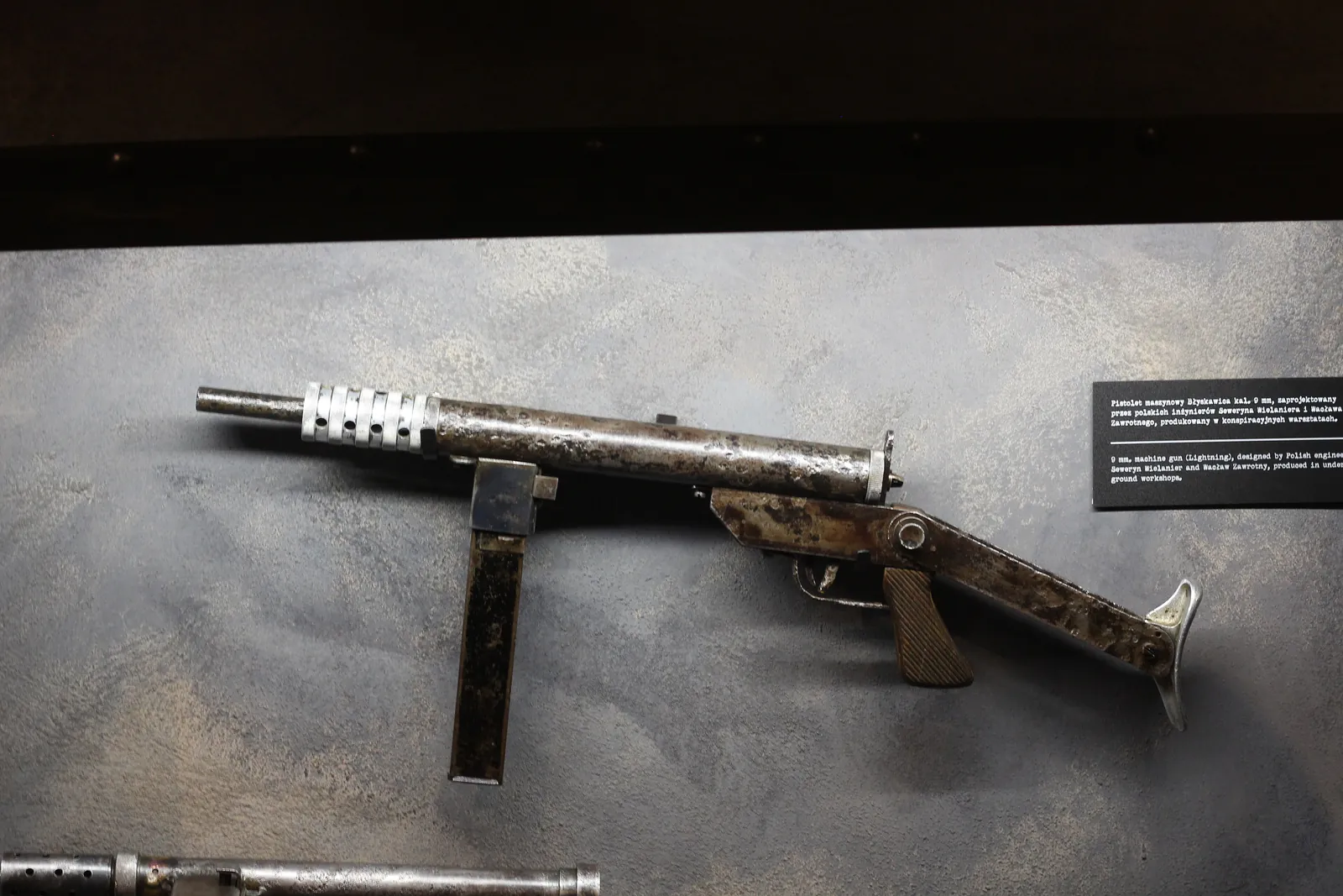 A slender tubular submachine gun with a perforated barrel jacket, a wooden foregrip and a wire stock, lying on a plain surface