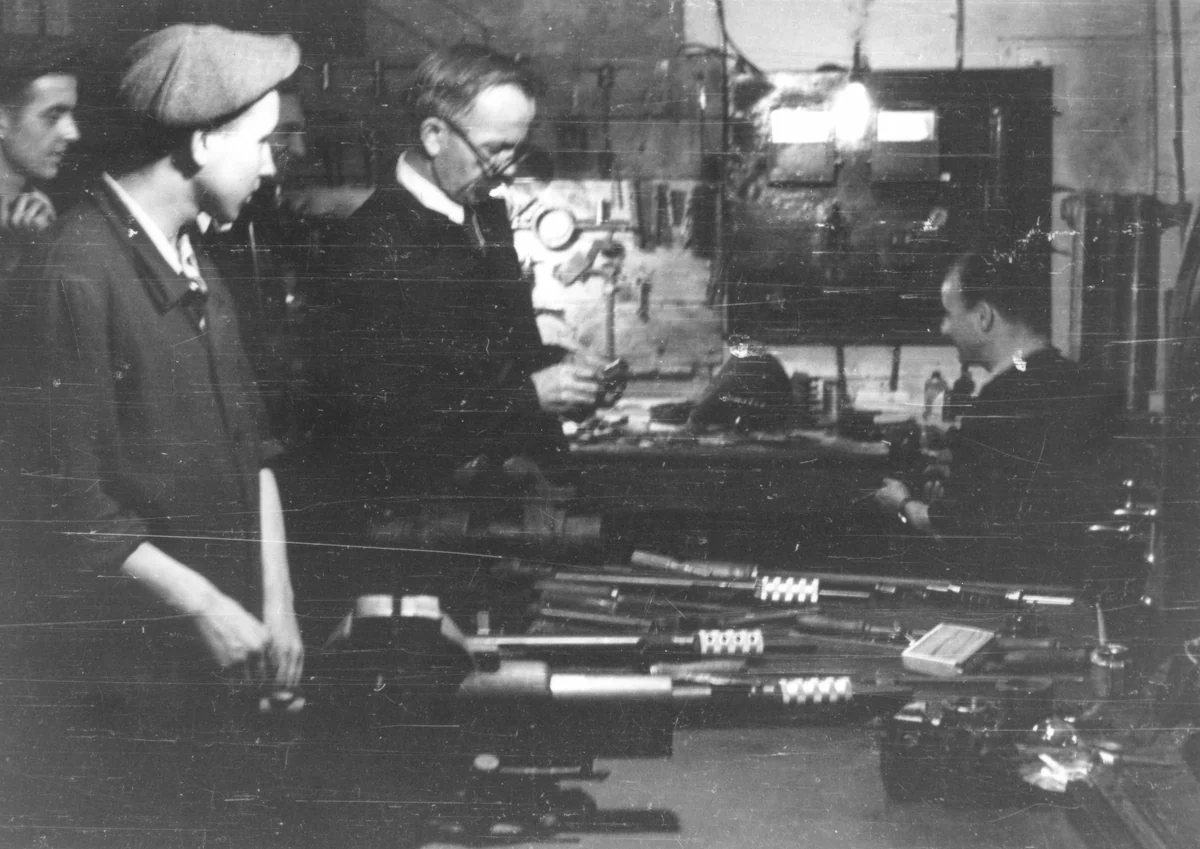 Men working at benches in a cramped clandestine workshop during the Warsaw Uprising, with submachine gun parts and tools visible on the table