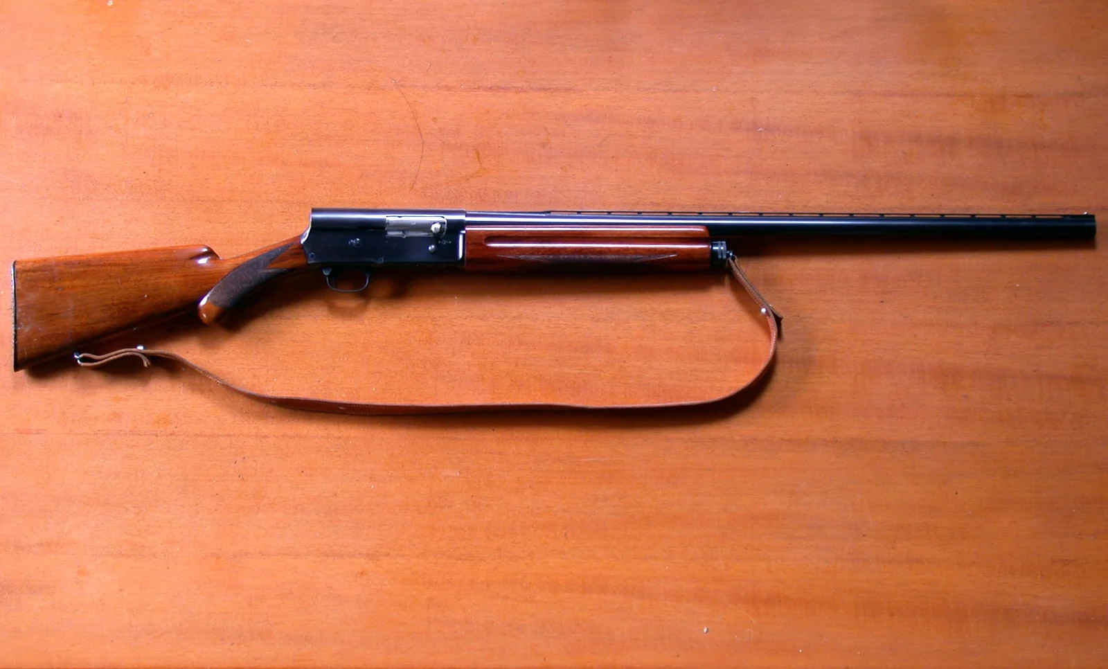 A Browning Auto-5 shotgun lying on an orange-brown wooden surface, seen from the left, with a leather sling, a walnut stock and forend, and a receiver squared off at the rear