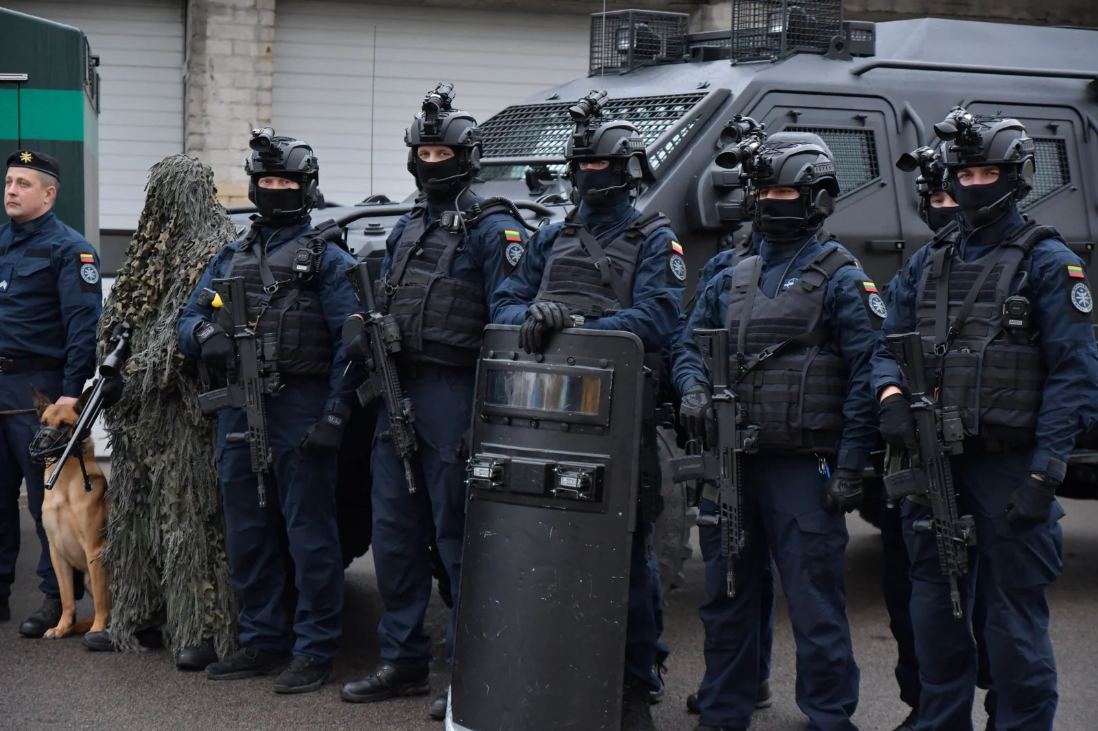 A line of police tactical officers in navy uniforms, black helmets with night-vision mounts and plate carriers, standing in front of a black armoured vehicle; several hold black short-barrelled rifles with vertical foregrips, and one holds a ballistic shield.