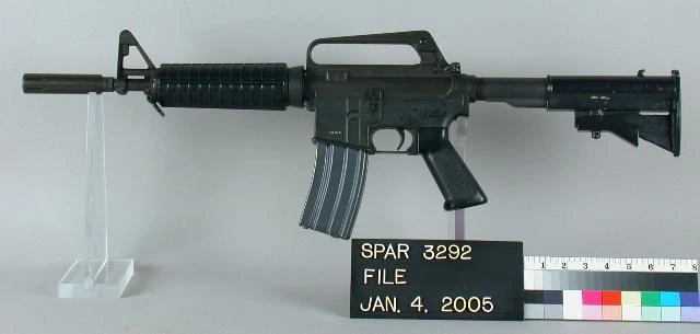 A short black carbine with a fixed carrying handle over the receiver, a ribbed handguard and a curved box magazine, resting on a museum display stand.