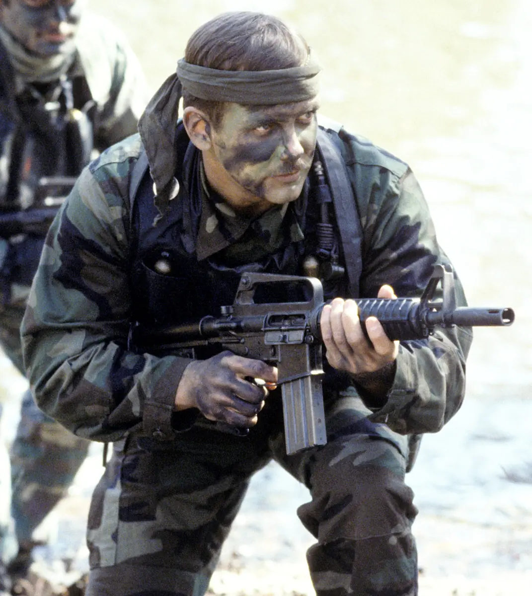 A sailor in camouflage kneels holding a short carbine with a magazine inserted, another sailor visible behind him during a field exercise.