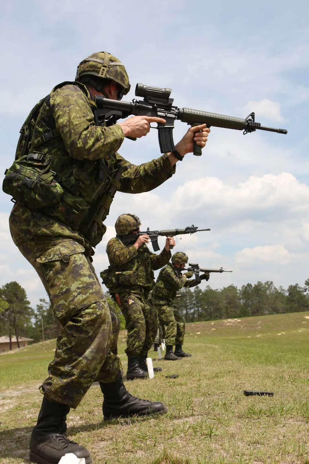 A line of soldiers in camouflage uniforms and helmets aiming black AR-15-pattern rifles fitted with carrying handles and optical sights, standing shoulder to shoulder on a firing line