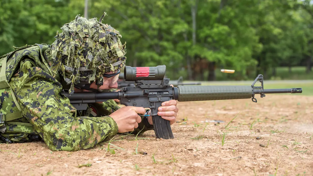 A soldier in camouflage aiming a black rifle fitted with a red-dot optical sight, with an ejected cartridge case visible in mid-air beside the ejection port