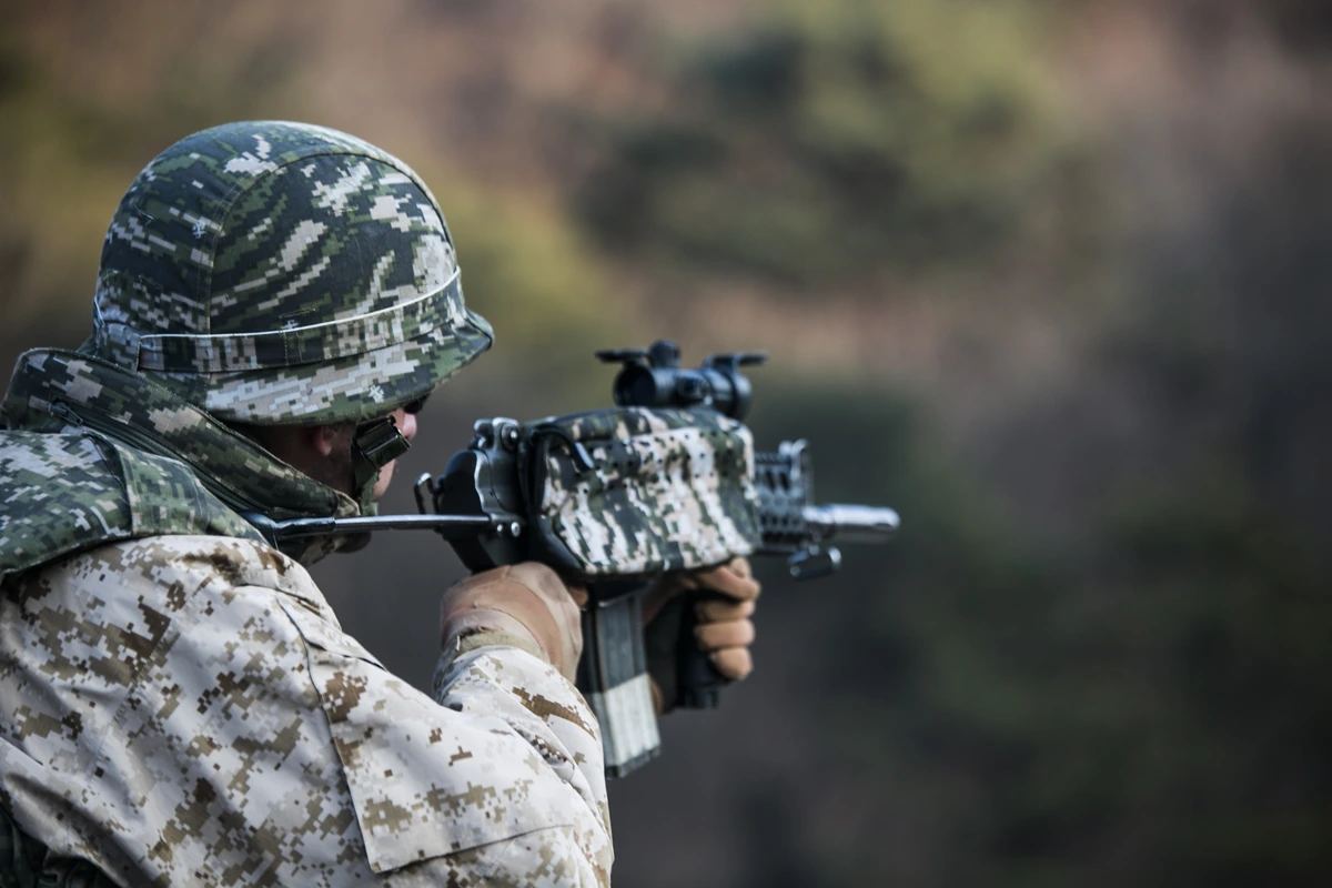 A Marine in digital camouflage fires a short carbine on a shooting range, another Marine visible behind him.