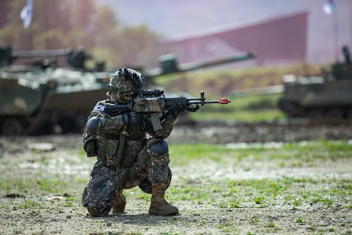 A kneeling soldier in digital camouflage aims a black rifle fitted with a quad-rail handguard and an optical sight, armoured vehicles visible out of focus behind him
