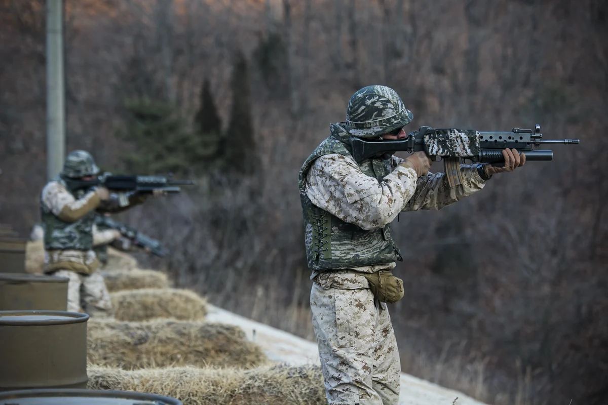 Two soldiers in camouflage fire black rifles fitted with underbarrel grenade launchers from a firing line of sandbags and drums