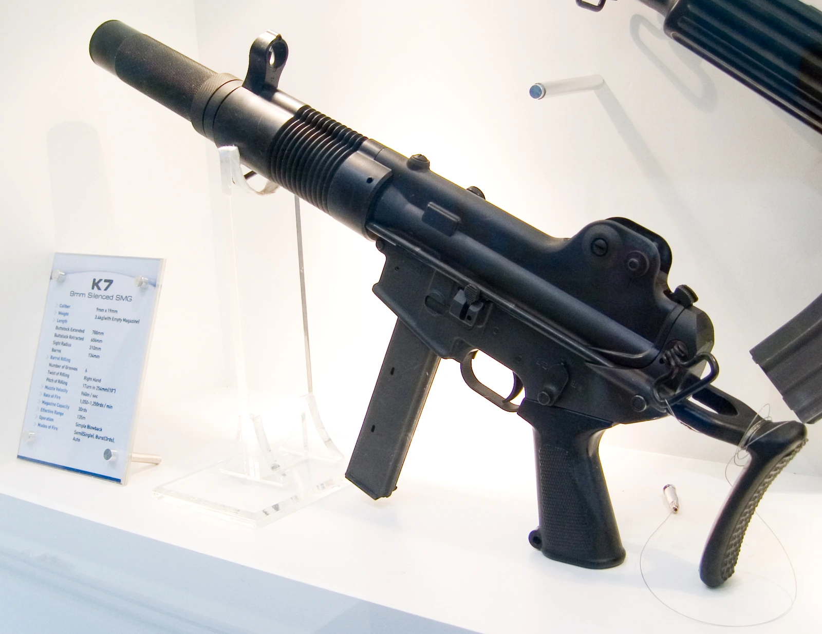A K7 submachine gun with a long ribbed suppressor housing and retractable stock, on a display stand