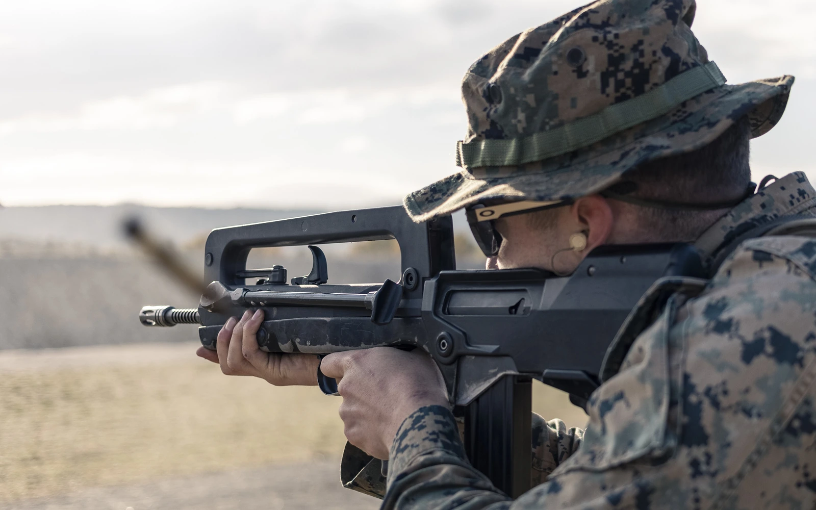 A US Marine in camouflage firing a FAMAS rifle at a range, seen over the shooter’s shoulder: a black polymer bullpup with a long carrying handle running most of its length