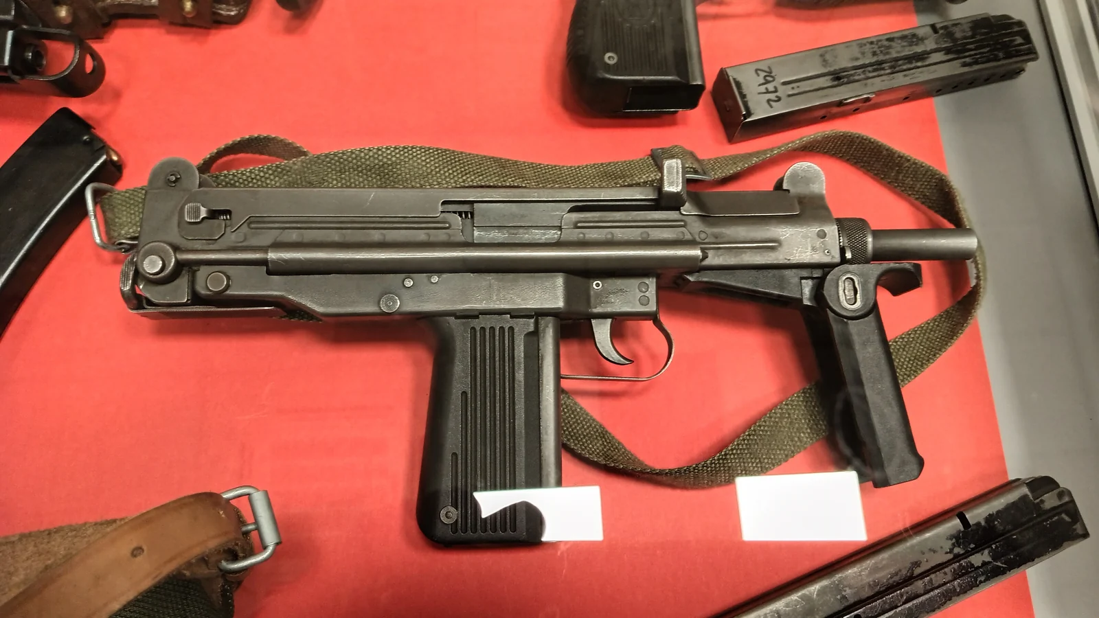 A dark, boxy submachine gun with a folding metal stock and a straight detachable magazine, displayed on a red cloth beside its holster and additional magazines