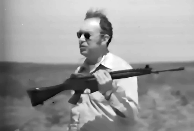 A man in civilian clothes holds an FN CAL rifle at port arms outdoors, demonstrating it to an unseen audience