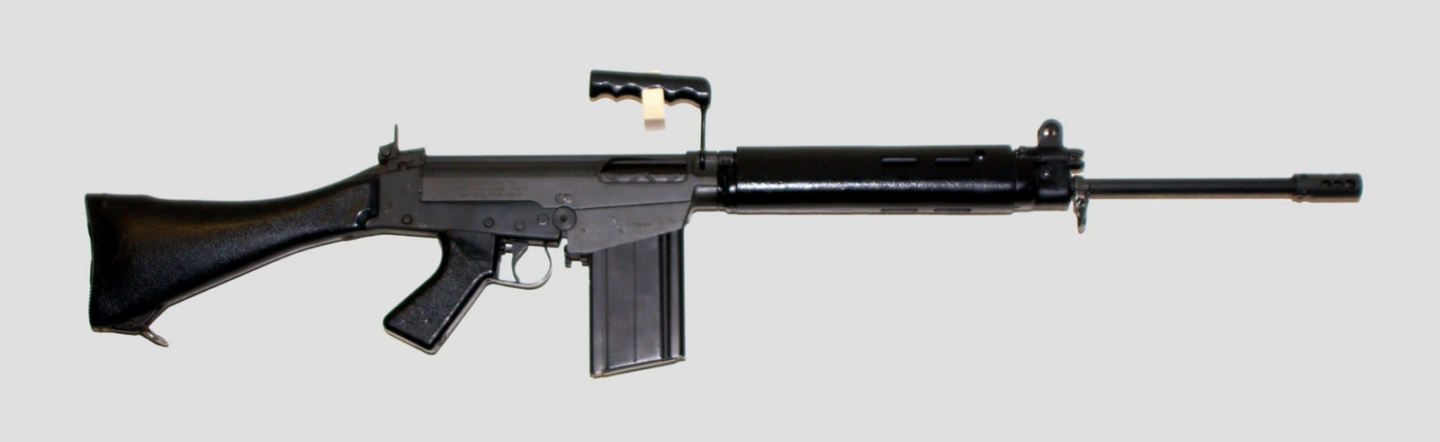 An FN FAL rifle isolated on a grey background, seen from the left, with a black synthetic stock, a carrying handle folded up over the receiver, a long handguard and a straight 20-round magazine