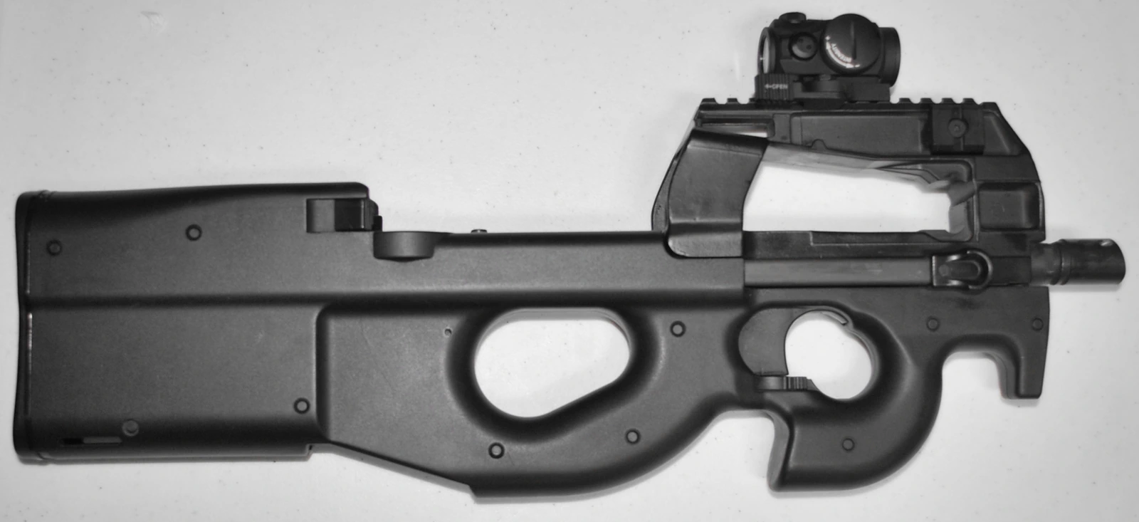 An FN P90 seen from the left against a pale background: a smooth polymer bullpup shell with a large thumbhole grip, a red-dot sight on top and a long translucent magazine lying flat along the receiver