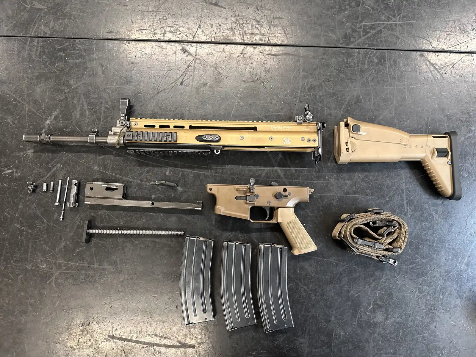 An FN SCAR-L disassembled on a steel workbench: tan upper receiver with the barrel installed, tan lower receiver, folding stock, bolt carrier, recoil spring, small parts, three magazines and a sling