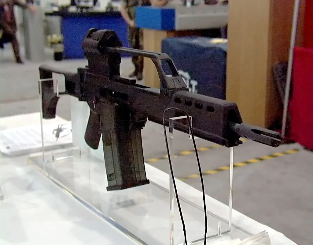A short black G36 carbine resting on clear acrylic stands on a white exhibition table, seen from the left. The stock is folded forward along the right side of the receiver, the handguard is short and slotted, and a bare muzzle with an open-pronged flash hider projects a little way past it.