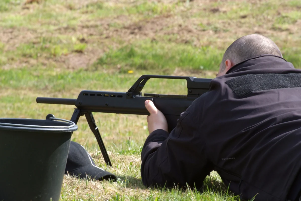 A man in a dark jacket lies prone on grass firing a G36 from a bipod, photographed from behind and to his left. The long slotted handguard, the open carry-handle sight bridge and the folded stock are visible in silhouette against the field.