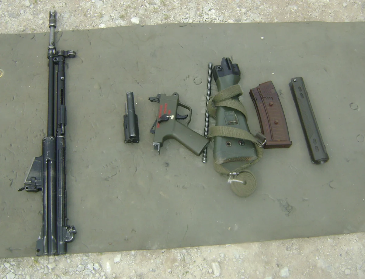 The barrel, bolt group, stock, pistol grip and magazine of a rifle laid out separately on a concrete surface.