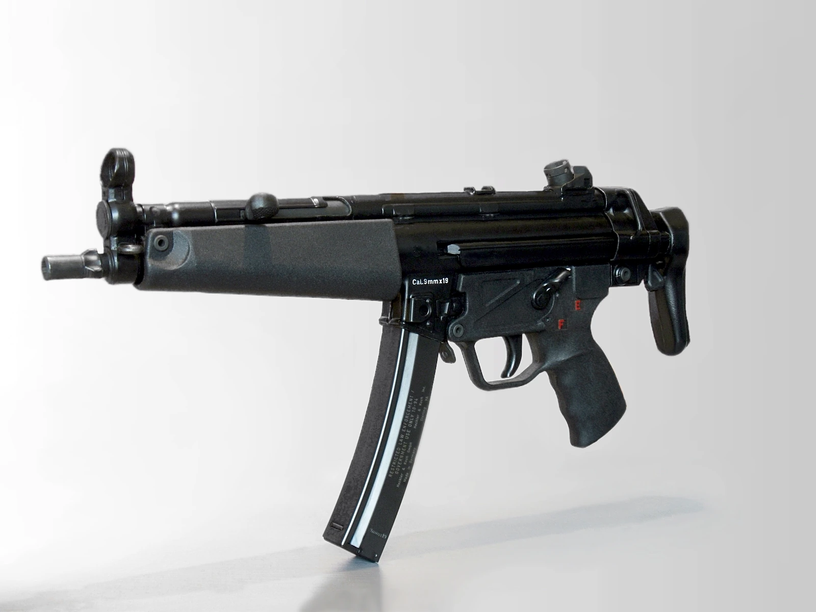 An MP5 submachine gun photographed at an angle against a white background, with a retractable stock, a curved magazine and “Cal. 9mm x 19” stamped on the receiver
