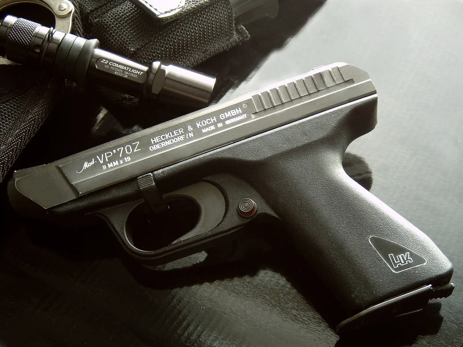 A black polymer-framed pistol lying on its side, slide markings reading 'Mod. VP70Z HECKLER & KOCH GMBH OBERNDORF/N MADE IN GERMANY 9 MM x 19', with a flashlight and holster in the background.