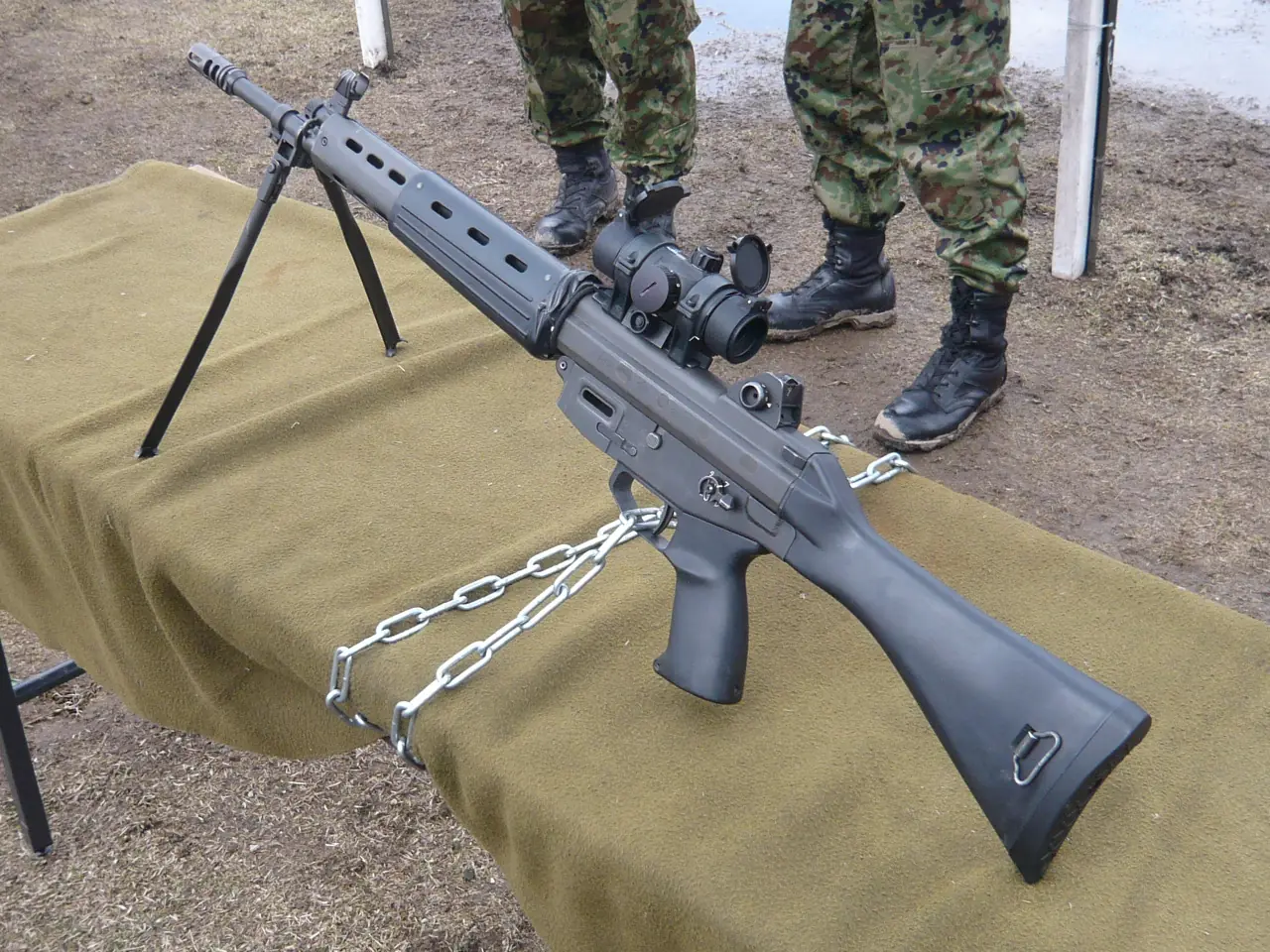 A black rifle with a fixed stock, a carrying handle over the receiver, a ribbed handguard and a mounted red-dot sight, laid on a table.