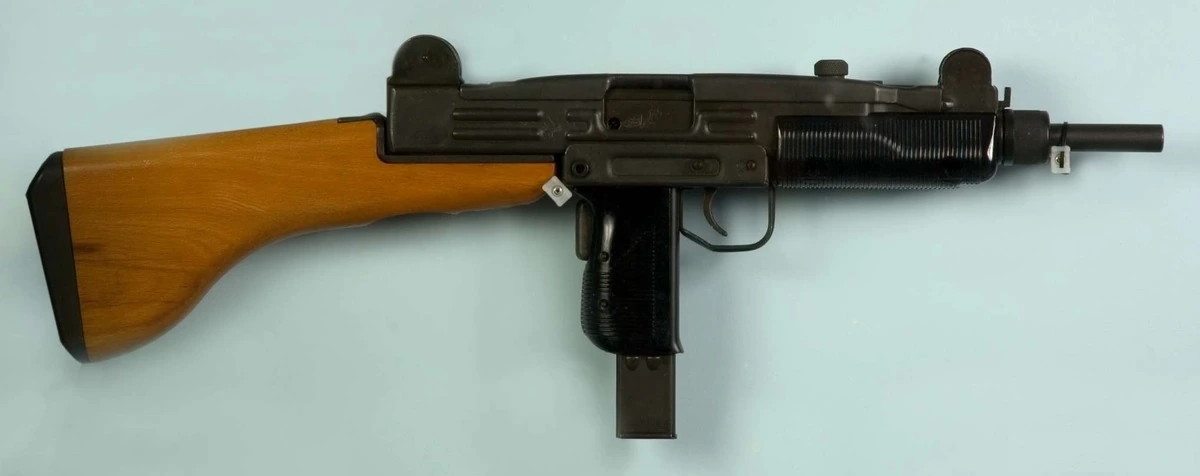 An Uzi submachine gun on a pale background, seen from the left, with a fixed wooden stock, a ribbed metal handguard and the magazine protruding from the pistol grip