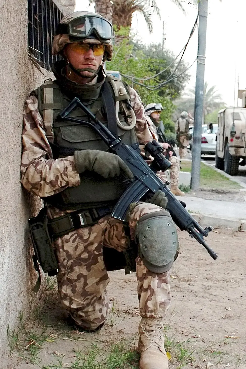 A soldier in desert camouflage crouching against a wall in an urban street, holding a Galil rifle with a folding tubular stock and a curved magazine