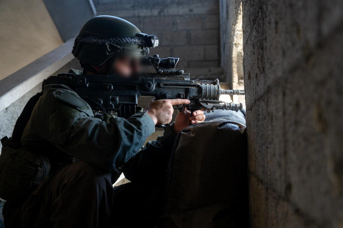 A helmeted soldier crouches in a doorway aiming a short bullpup rifle fitted with a magnified optic, resting the rifle on a sandbag.