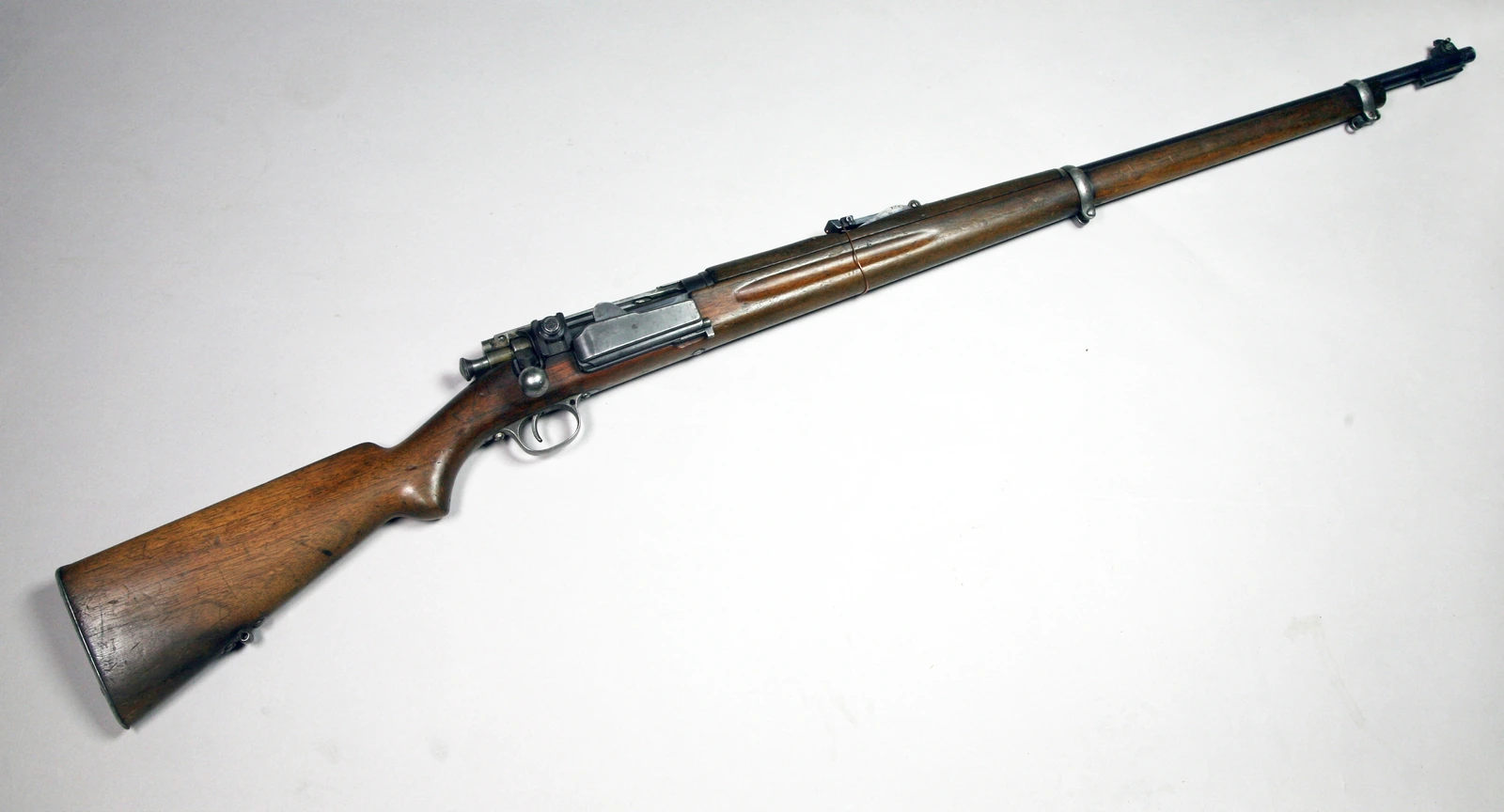 A bolt-action rifle photographed from the right against a pale grey ground, muzzle to the upper right. The stock is plain walnut without a pistol grip; a flat rectangular steel plate on the right of the receiver is the hinged door of the side magazine, and the bolt handle is a straight stem with a round knob. A tangent rear sight sits on the barrel well forward of the receiver, and the barrel is exposed for its last third with two bands.