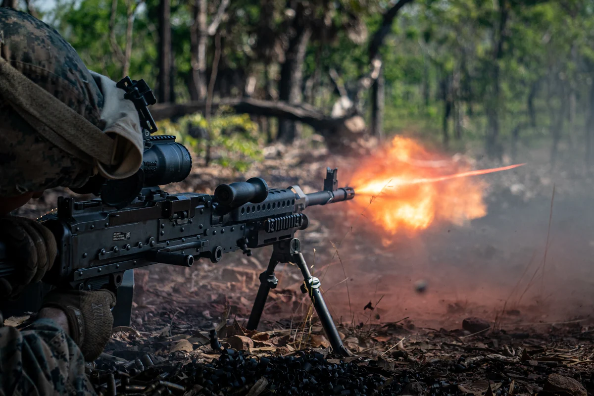 A soldier lying prone in a forest clearing firing a belt-fed machine gun on a bipod, with a burst of muzzle flash visible at the barrel