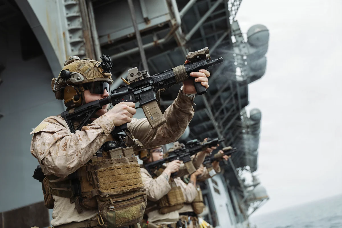 A line of Marines in desert camouflage firing rifles upward from the deck of a ship, the nearest one holding an M27 by a magwell grip with a red-dot sight on the rail and smoke drifting from the muzzle. The ship's superstructure and grey sea fill the background.
