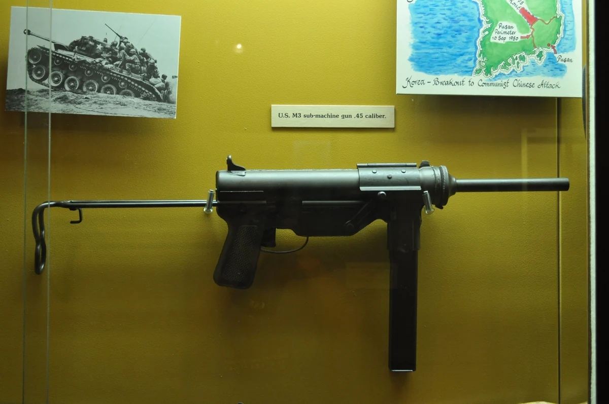 An M3 submachine gun mounted behind glass in a museum case against an ochre wall, with a printed label reading “U.S. M3 sub-machine gun .45 caliber”, a photograph of a tank crew above it and a map of Korea to the right.