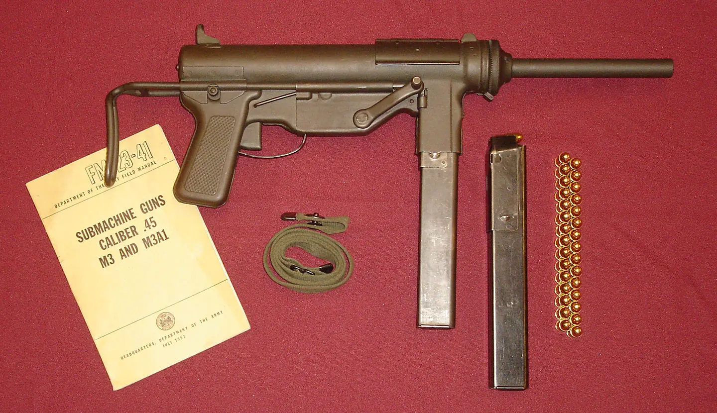 An M3 submachine gun laid on red cloth beside its field manual, a canvas sling, a spare magazine and thirty loose cartridges. The gun is a plain steel tube with a pistol grip, a straight box magazine below it and a bent wire stock extended to the rear.
