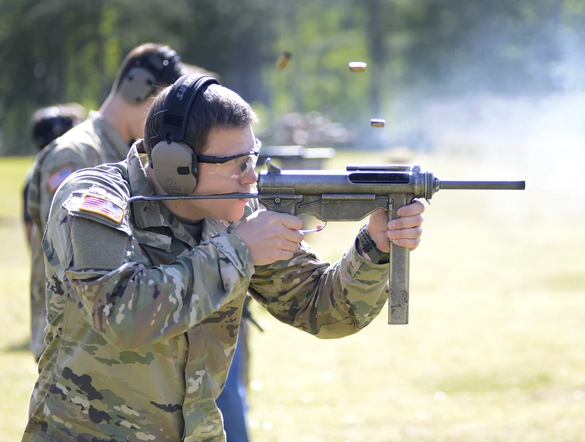 A soldier in camouflage fires an M3A1 submachine gun one-handed on an outdoor range, spent cases in the air beside the ejection port and smoke drifting from the muzzle.