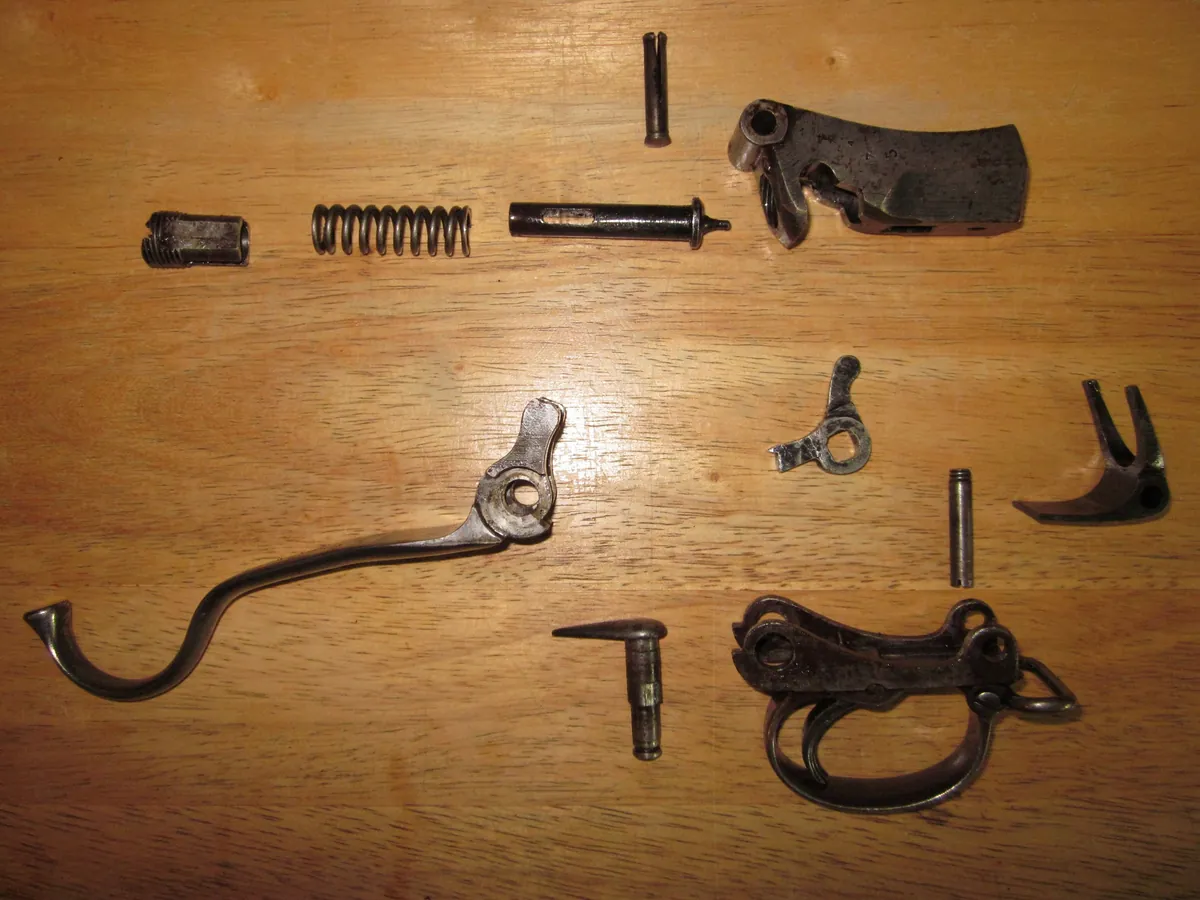 The disassembled falling-block action of a Martini-Henry Mark II laid out on a wooden bench: the breech block, hammerless striker and coil spring, the underlever, the tumbler and sear, and assorted pins