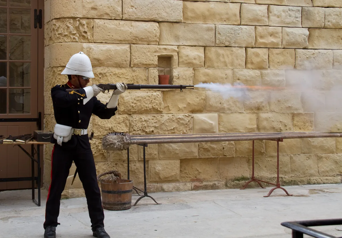 A re-enactor in Victorian British Army uniform and pith helmet firing a Martini-Henry rifle from a bench rest against a stone wall, smoke and muzzle flash visible at the muzzle