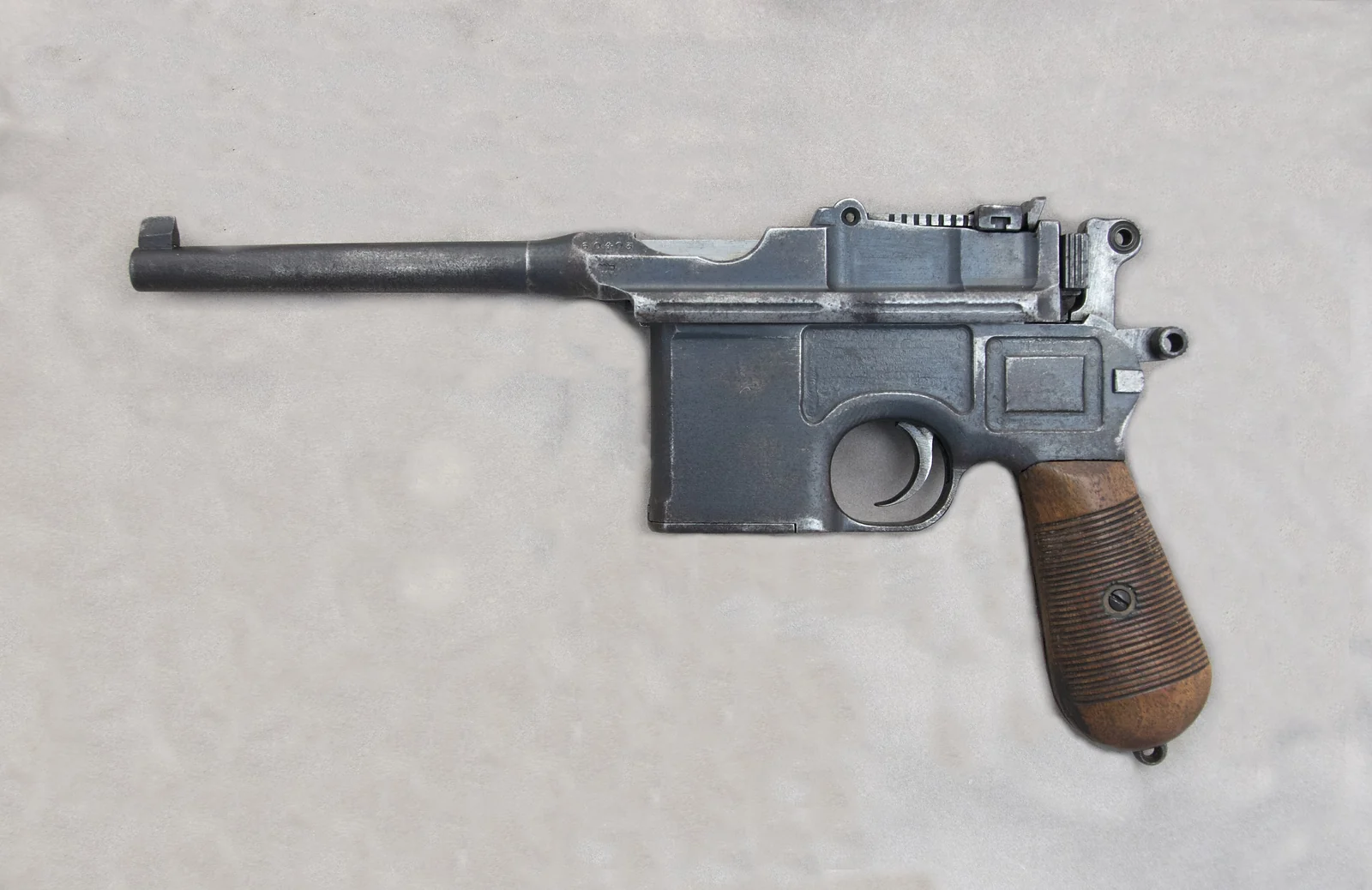 A blued Mauser C96 pistol seen from the left, showing its long barrel, fixed magazine ahead of the trigger guard, sweeping wooden grip and a rear tangent sight on top of the barrel