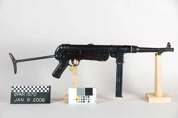 An MP 40 submachine gun photographed against a grey museum background with a colour calibration chart, stock folded under, the magazine hanging vertically