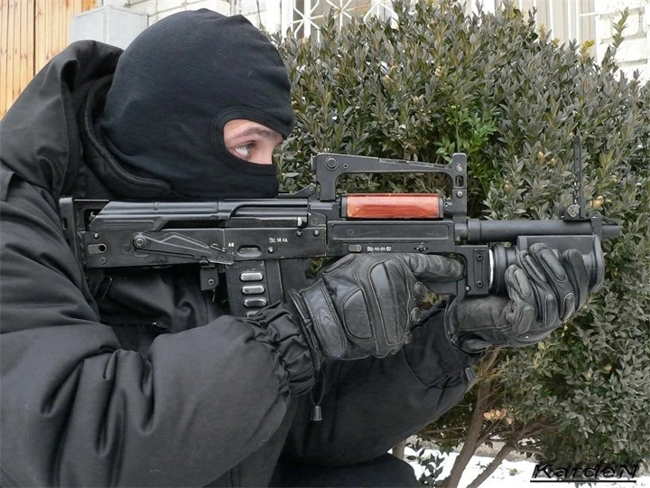 A man in a black balaclava, black winter jacket and black gloves crouching in snow beside a hedge, shouldering a bullpup weapon with a wooden-coloured handguard visible under the carrying handle and a wide tube under the barrel. The photograph carries a small watermark in the corner.