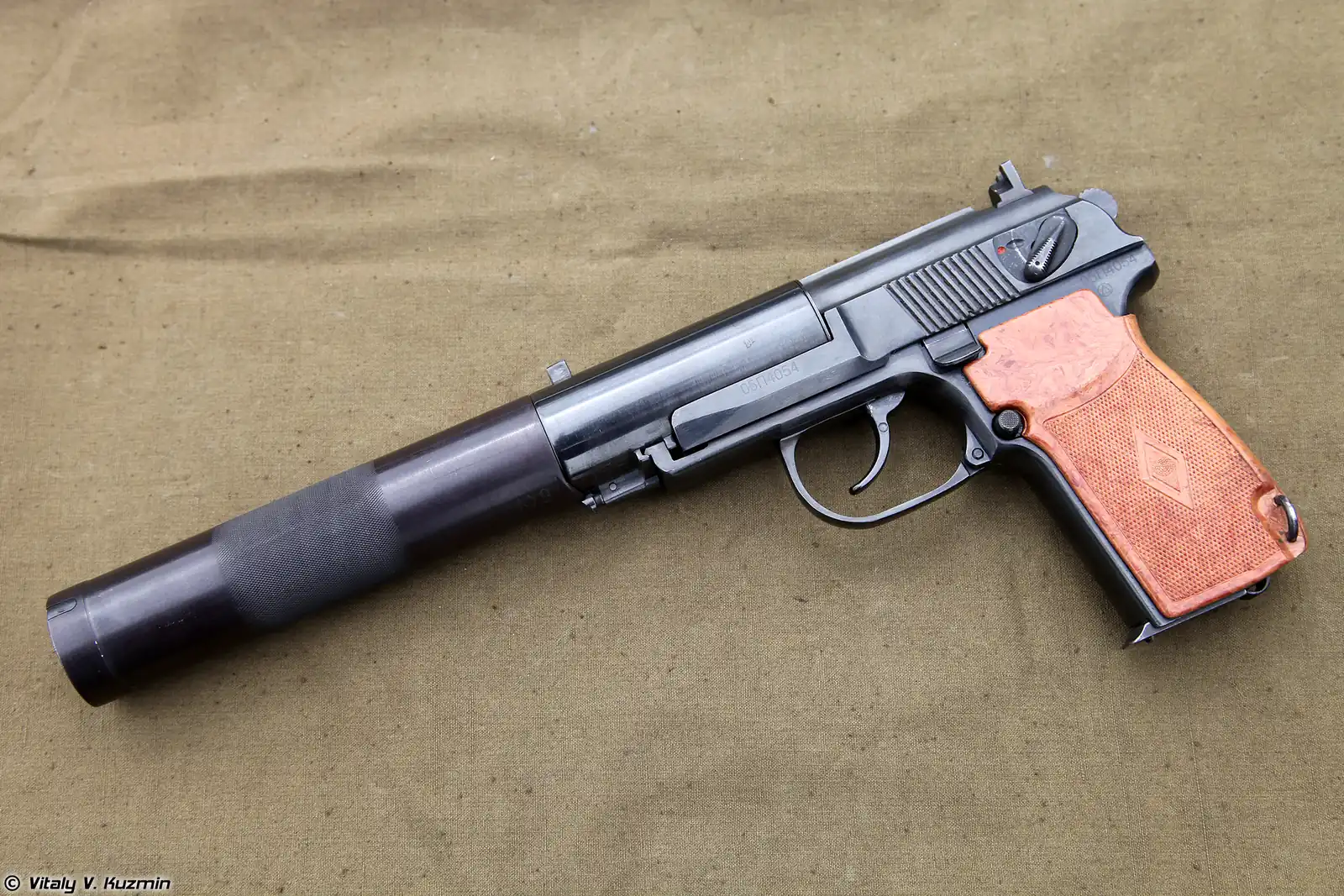 A blued semi-automatic pistol with a fixed cylindrical suppressor sleeve over its barrel and wooden grip panels, photographed against a canvas background