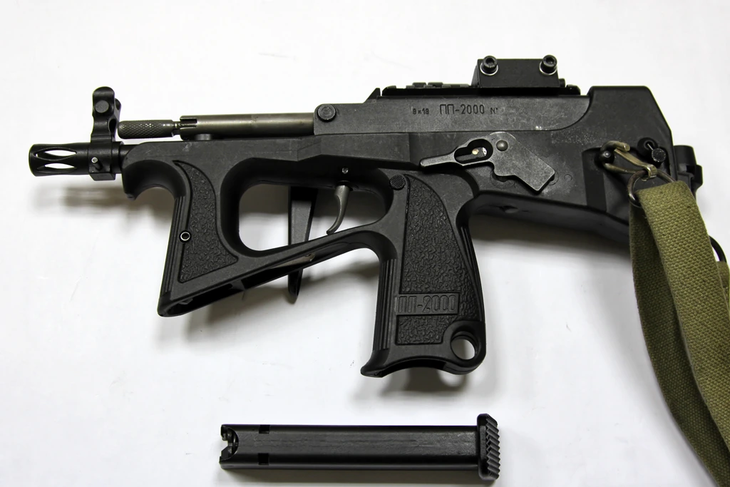 A PP-2000 submachine gun lying on a white surface with its box magazine removed and set below the grip, showing the open magazine well in the pistol grip.