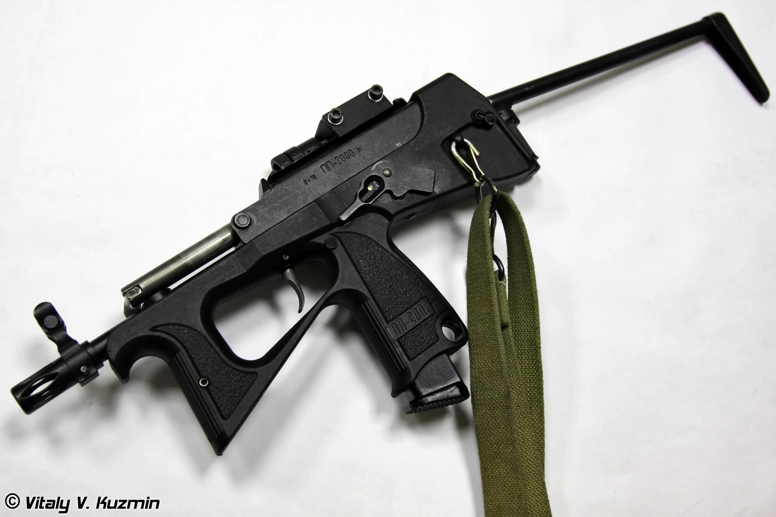A compact black bullpup-style submachine gun lying on a white surface, with a wire skeleton stock folded back along the top of the receiver, a triangular integral trigger guard doubling as a foregrip, and a green sling attached at the rear.