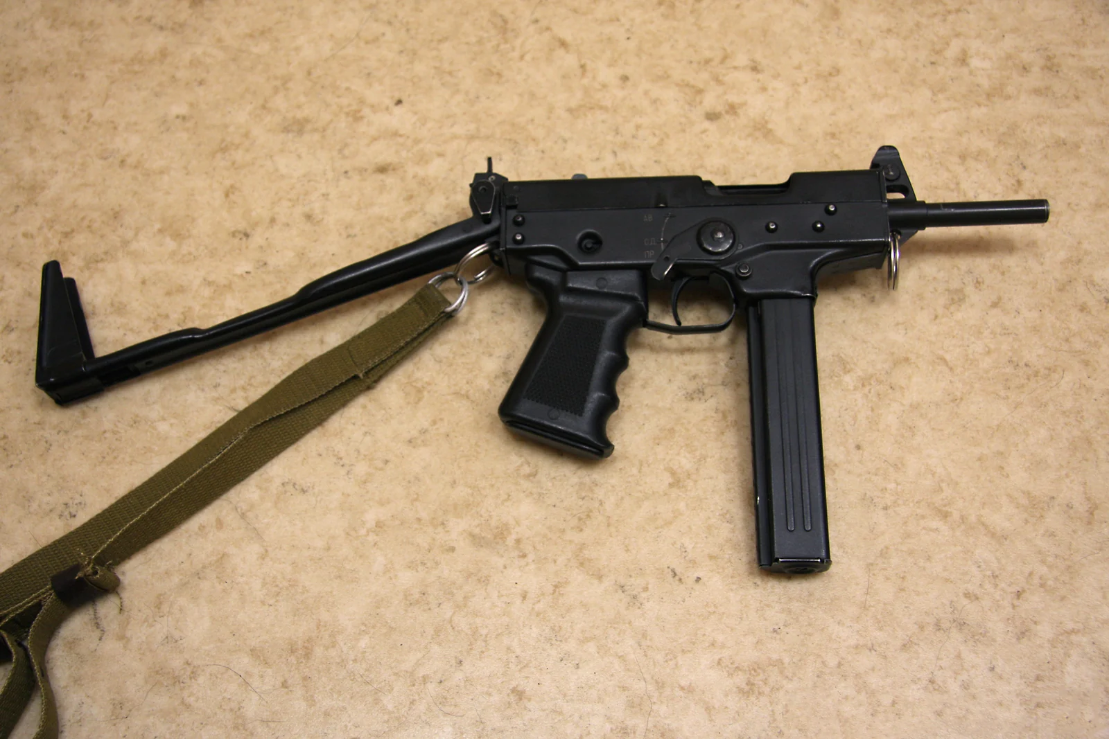 A compact black submachine gun with a folded wire stock, a wide pistol grip and a curved magazine, lying on a tan surface with its sling